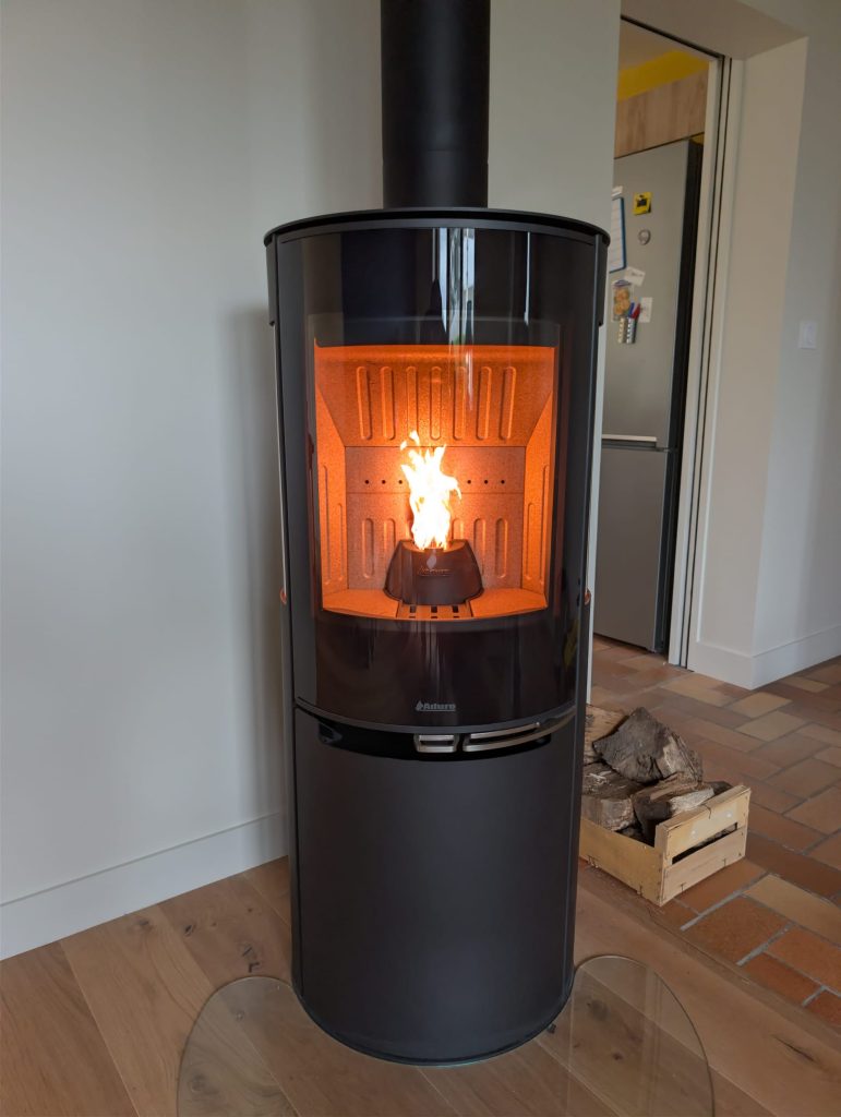 Poêle mixte bois et granulés Aduro, cylindre noir posé sur une plaque de sol en verre, à côté d’une caisse de bûches